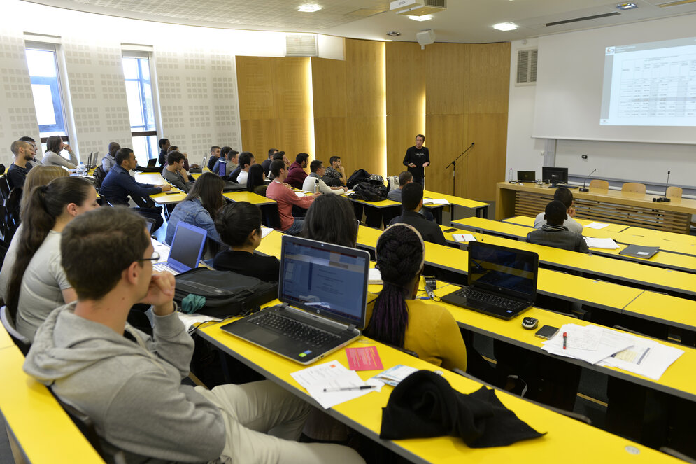 Étudiants en amphithéâtre suivant un cours avec ordinateurs portables, formateur devant un écran de projection